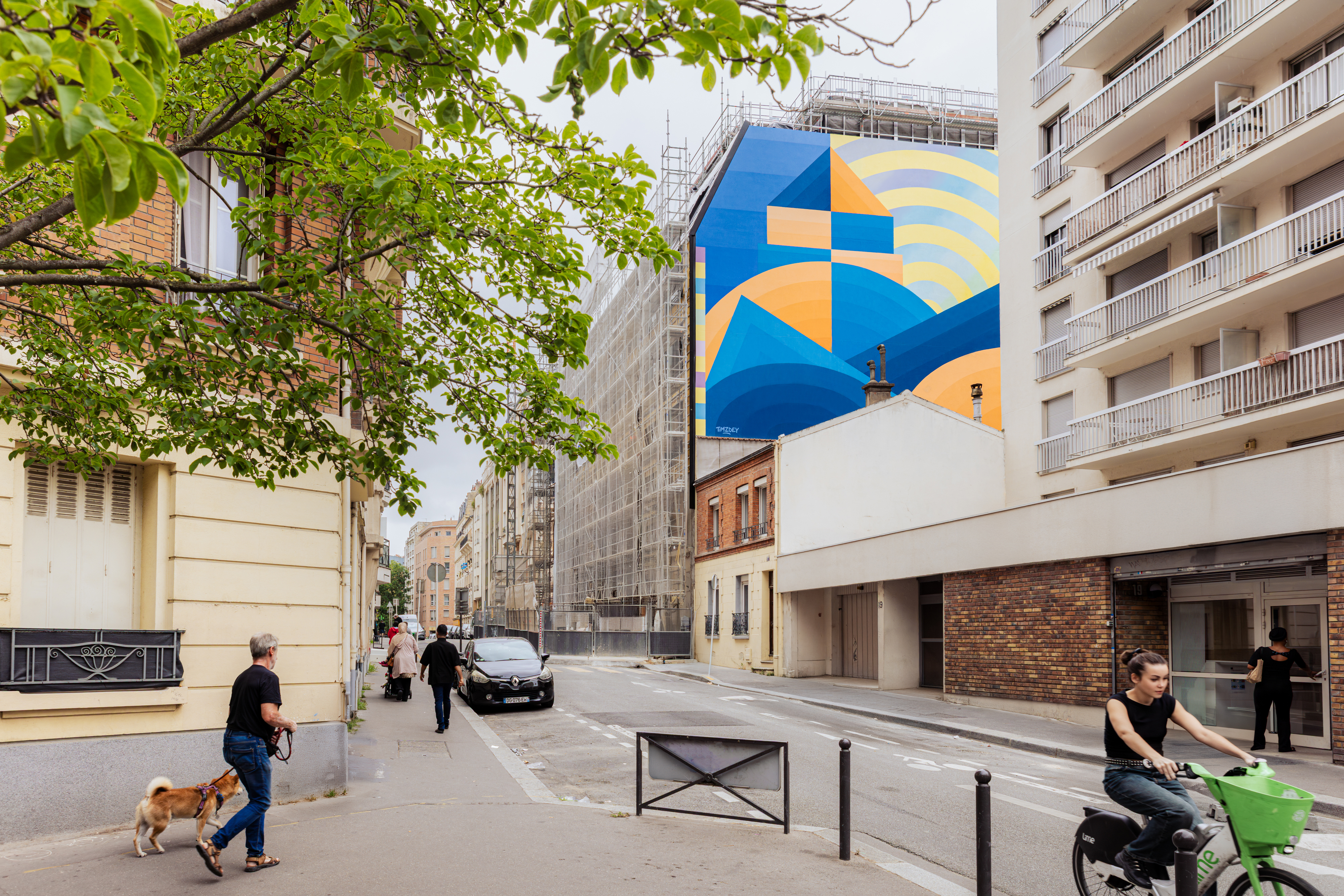 Fresque sur un mur avec des formes géométrique ronde et triangulaire de couleurs bleu et orange et une maison dessinée qui représente le dôme de la Sorbonne. Dans la rue, une fille brune est à vélo, un monsieur promène son chien (shiba) et une dame rentre dans un building
