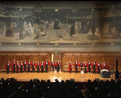 Photos de loin d'une dizaine de professeurs alignées face à la scène et au public en toge rouge avec la fresque du Grand Amhithéâtre de la Sorbonne derrière eux