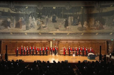 Photos de loin d'une dizaine de professeurs alignées face à la scène et au public en toge rouge avec la fresque du Grand Amhithéâtre de la Sorbonne derrière eux