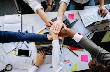 Photo avec une prise de vue du dessus avec 5 mains qui se joignent au-dessus d'un bureau dont une d'un homme de 50 ans au cheveux blanc et une main sur un ordinateur
