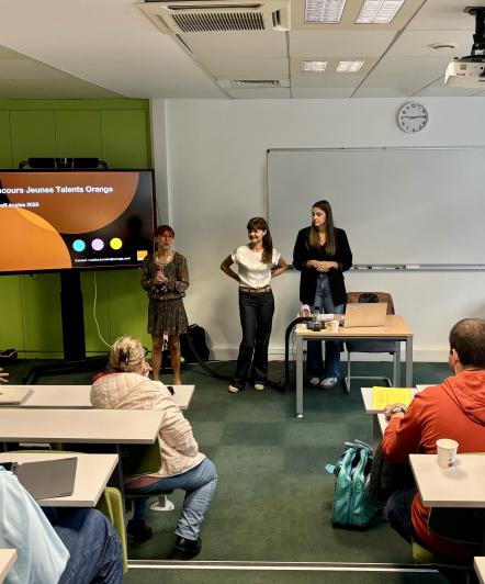 Amphithéâtre avec des élèves pris de dos et deux personnes qui présente sur un écran le Concours Jeunes Talents d'Orange. 