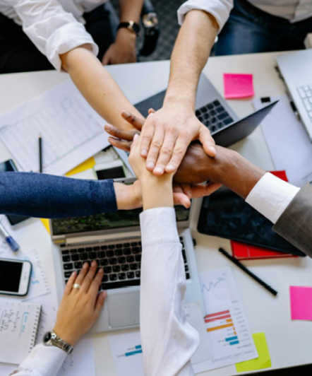 Photo avec une prise de vue du dessus avec 5 mains qui se joignent au-dessus d'un bureau dont une d'un homme de 50 ans au cheveux blanc et une main sur un ordinateur