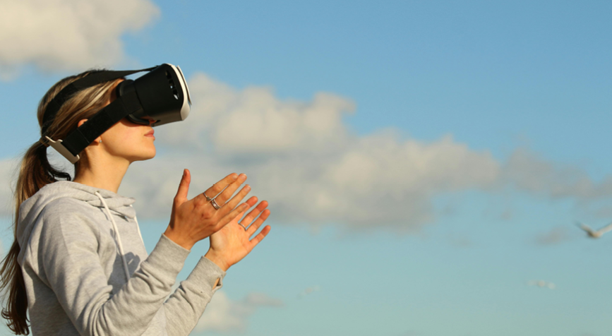 Une jeune femme blonde avec un casque de réalité virtuelle de profil et un sweat gris que l'on voit de profil. En fond, des nuages et un ciel bleu