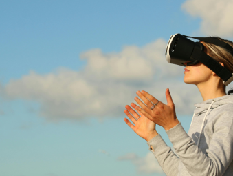 Une jeune femme blonde avec un casque de réalité virtuelle de profil et un sweat gris que l'on voit de profil. En fond, des nuages et un ciel bleu 