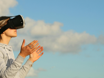Une jeune femme blonde avec un casque de réalité virtuelle de profil et un sweat gris que l'on voit de profil. En fond, des nuages et un ciel bleu