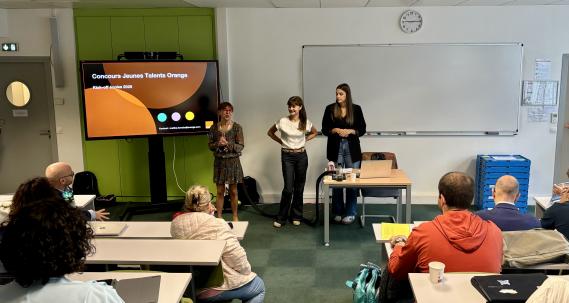 Amphithéâtre avec des élèves pris de dos et deux personnes qui présente sur un écran le Concours Jeunes Talents d'Orange. 