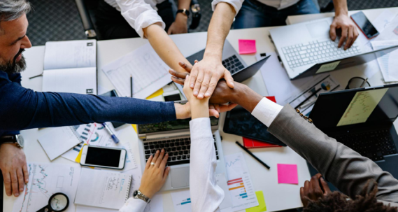 Photo avec une prise de vue du dessus avec 5 mains qui se joignent au-dessus d'un bureau dont une d'un homme de 50 ans au cheveux blanc et une main sur un ordinateur